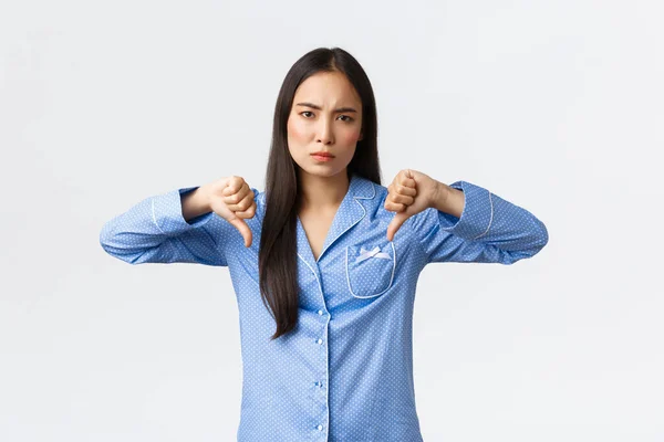 Decepcionado frunciendo el ceño mujer asiática en pijama azul expresan aversión, mostrando pulgares hacia abajo escéptico, juzgando mal producto o cosméticos para el cuidado de la piel, siendo disgustado con la calidad, fondo blanco — Foto de Stock