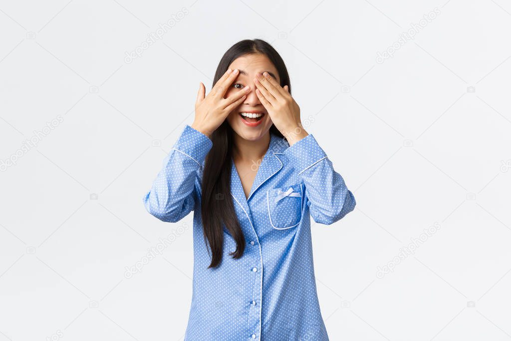Cheerful smiling asian girl in blue pajamas waking up to surprise gift on birthday, wearing jammies, close eyes and peeking through fingers amazed, looking curious at something, white background