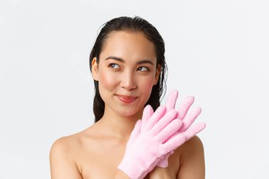 Kişisel bakım, kadın güzelliği, banyo ve duş konsepti. Beyaz arka planda duran güzel, çıplak Asyalı kadının yakın çekimi, sol düşünceli, banyo eldiveni kullanırken kurnazca gülümseyen, ellerini ovuşturan