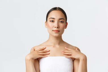 Temiz ciltli, mükemmel yüzlü, havlunun içinde duran, vücuduna dokunan, krem ya da temizleyici süren, banyodaki cilt bakımı ürünleri yapan, spa salonuna giden, beyaz arka planda yaşayan güzel Asyalı bir kadın.