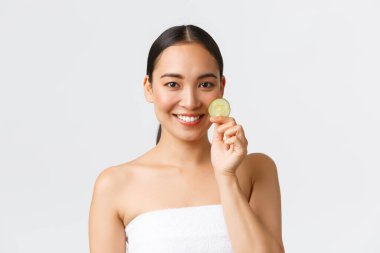 Güzellik, kişisel bakım, kaplıca salonu ve cilt bakımı konsepti. Banyo havlusu giymiş, gülümseyen ve elinde salatalık parçası olan güzel bir Asyalı kadın. Temizleyici jel, yüz maskesi, nemlendirici veya duş için vücut jeli.