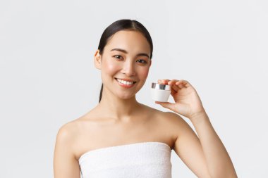 Güzellik, kişisel bakım, kaplıca salonu ve cilt bakımı konsepti. Banyo havlusu içinde gülümseyen güzel Asyalı kız. Krem ve gülümseme gösteriyor. Kozmetik ürünler öneriyor.