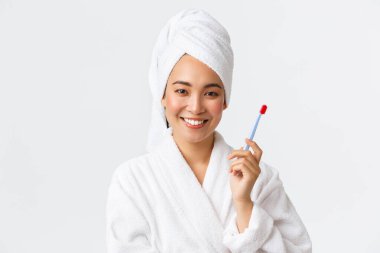 Kişisel bakım, kadın güzelliği, banyo ve duş konsepti. Yüzünde bornoz ve havlu olan, saçını yıkayan, diş fırçasıyla dişlerini fırçalayan, beyaz arka planda duran çekici Asyalı kadının yakın çekimi.