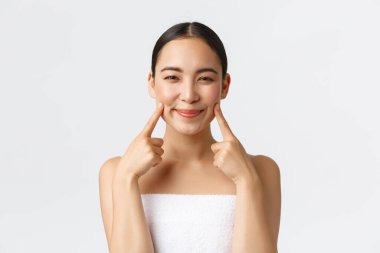 Güzellik, kozmetoloji ve kaplıca salonu konsepti. Yanaklarını gösteren, gülümseyen, cilt bakımı yapan, temizleyici öneren, beyaz arkaplan öneren Asyalı güzel kızın yakın çekimi.