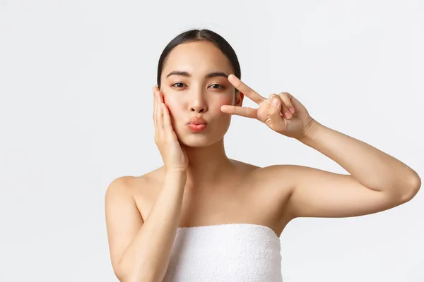 Beauty, personal care, spa salon and skincare concept. Close-up of sensual pretty asian girl in bath towel showing peace kawaii gesture and blowing air kiss, satisfied with skin condition — Stock Photo, Image