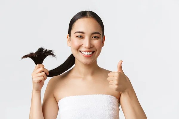 Kecantikan, produk rambut rontok, sampo dan konsep perawatan rambut. Close-up of satisited, happy asian girl in bath towns shows thumbs-up and healthy hair ends, standing pleased white background — Stok Foto