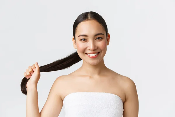 Kecantikan, produk rambut rontok, sampo dan konsep perawatan rambut. Close-up of beautiful asian woman in bath handuk shows strong and healthy hair, holding ponytail and smile happy, white background — Stok Foto