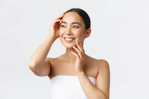 Bella ragazza asiatica tenera in asciugamano guardando lontano soddisfatto, toccando viso pulito, senza acne, godendo la giornata in spa, in piedi bagno su sfondo bianco, applicare detergente o prodotto per la cura della pelle — Foto Stock