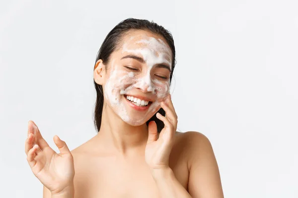 Cuidado de la piel, belleza femenina, higiene y concepto de cuidado personal. Primer plano de hermosa chica coreana sonriente cerrar los ojos y mirando encantado mientras se utiliza espuma limpiadora para lavar el maquillaje, fondo blanco — Foto de Stock