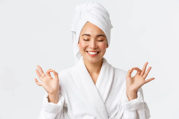 Cuidados pessoais, mulheres beleza, banho e chuveiro conceito. Close-up de aliviado atraente mulher asiática assistir spa salão de beleza, use toalha de banho e roupão de banho, meditando, sentindo nirvana, sorrindo — Fotografia de Stock