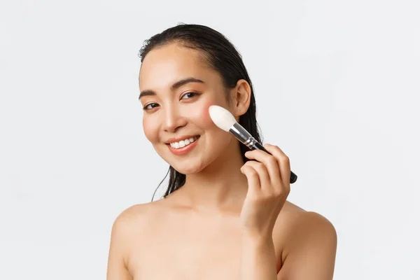 Skincare, kecantikan wanita, kebersihan dan konsep perawatan pribadi. Close-up dari wanita Asia muda yang cantik menggunakan makeup kuas di pipi dan tersenyum, berdiri telanjang di atas latar belakang putih — Stok Foto