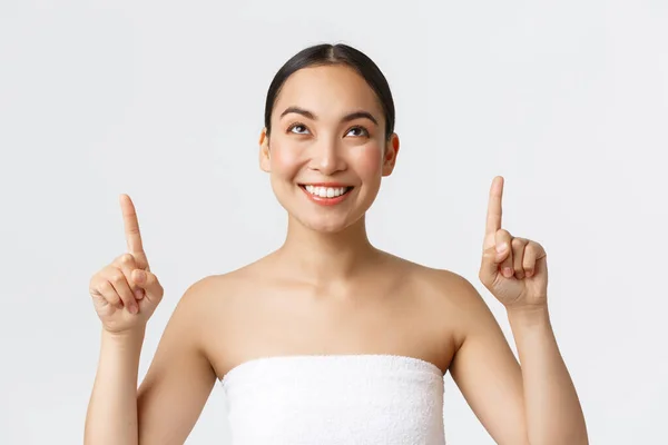 Beauty, cosmetology and spa salon concept. Beautiful curious asian woman in towel seeing interesting news, pointing fingers up and looking top with pleased smile, white background — Stock Photo, Image