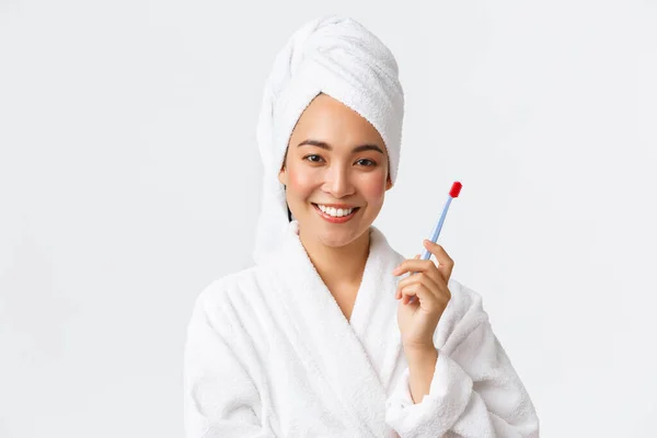 Personlig vård, kvinnlig skönhet, bad och dusch koncept. Närbild av leende attraktiv asiatisk kvinna bär badrock och handduk, tvättat hår, borsta tänderna med tandborste, stående vit bakgrund — Stockfoto