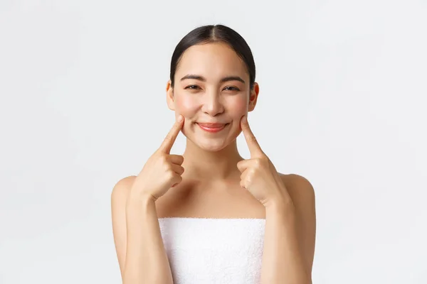Kecantikan, kosmetologi dan konsep salon spa. Close-up dari gadis Asia konyol yang cantik dengan handuk mandi menunjuk pipinya dan tersenyum bahagia, menerapkan produk perawatan kulit, merekomendasikan bersih, latar belakang putih — Stok Foto
