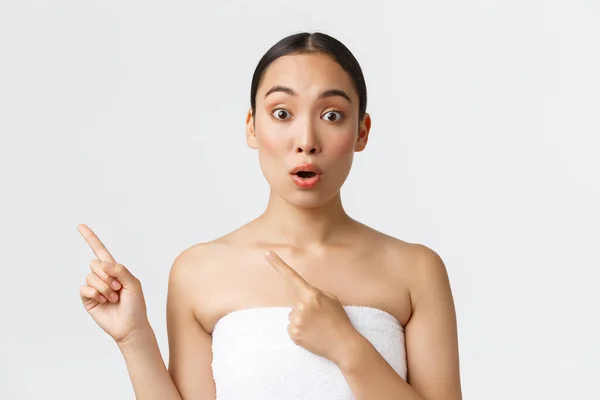 Amazed and impressed beautiful asian woman in towel pointing fingers upper left corner and looking fascinated, recommend cleanser, skincare product, spa salon services, white background — Stock Photo, Image