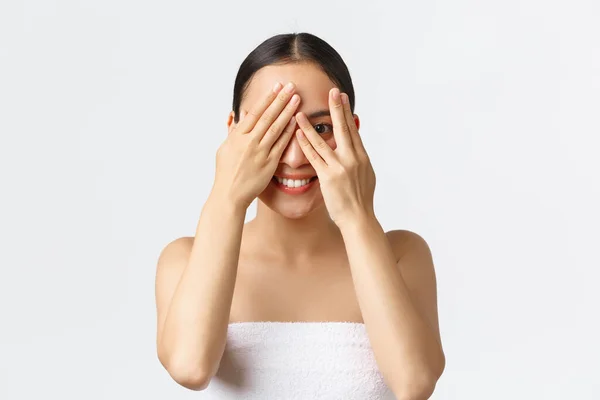 Beleza, cosmetologia e conceito de salão de spa. Silly bela menina asiática na toalha de banho espreitando através dos dedos como olhos de cobertura com as mãos e sorrindo, interessado que surpresa ela exceto, fundo branco — Fotografia de Stock