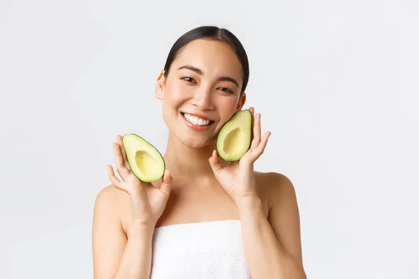 Beauté, soins personnels, spa et concept de soins de la peau. tendres mignon asiatique femelle dans serviette de bain montrant avocat et souriant satisfait, nourrir et traiter la peau avec des masques pour le visage ou crème, fond blanc — Photo