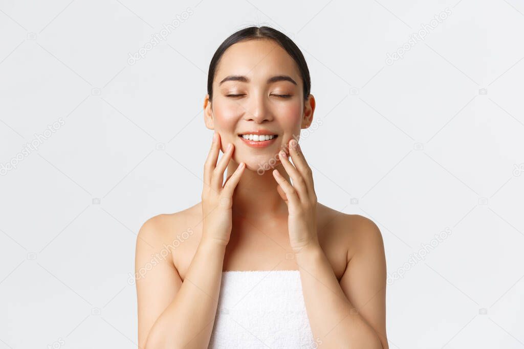Tender smiling beautiful asian girl attend spa salon, standing in towel, close eyse and touching soft clean skin, satisfied with cream or cleansing gel results, standing white background