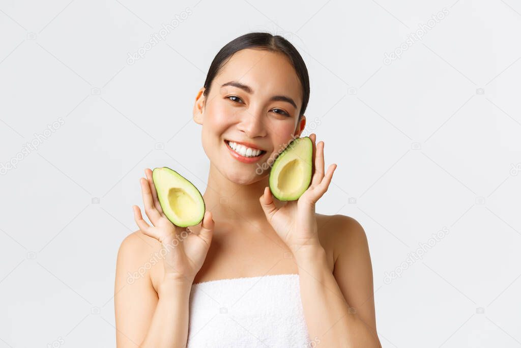 Beauty, personal care, spa and skincare concept. Tender cute asian female in bath towel showing avocado and smiling satisfied, nourish and treat skin with face masks or cream, white background