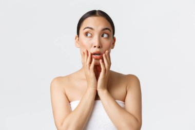 Güzellik, kozmetoloji ve kaplıca salonu konsepti. Havlulu güzel Asyalı kız şaşırmış ve şok olmuş sol üst köşede, temiz bir yüze dokunuyor, beyaz arka planda duruyor.