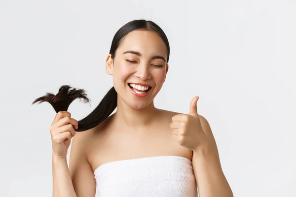 Kecantikan, produk rambut rontok, sampo dan konsep perawatan rambut. Close-up of satisited, happy asian girl in bath towns shows thumbs-up and healthy hair ends, standing pleased white background — Stok Foto