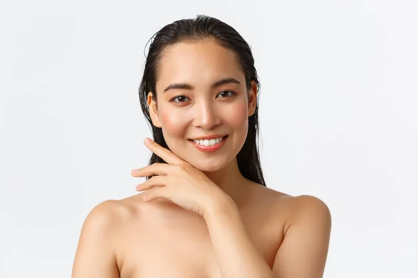 Skincare, bodycare, beauty and bath concept. Close-up of sensual attractive asian woman standing naked in shower, smiling and gently touching face, promo of facial products, white background — Stock Photo, Image