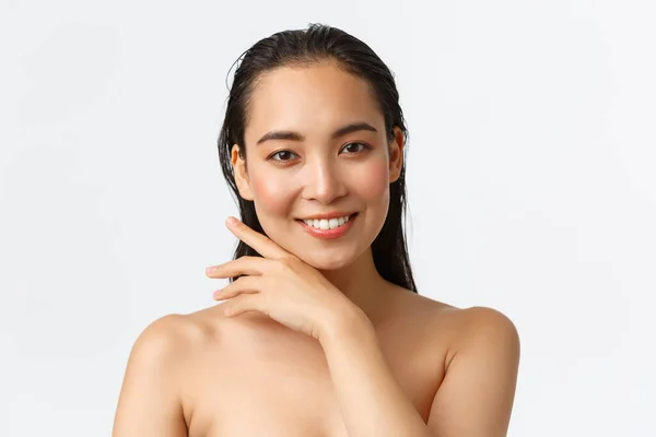 Cura della pelle, cura del corpo, bellezza e concetto di bagno. Primo piano di sensuale attraente donna asiatica in piedi nuda sotto la doccia, sorridente e delicatamente toccante viso, promo di prodotti per il viso, sfondo bianco — Foto Stock