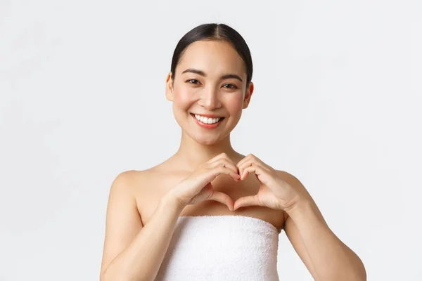 Bellezza, cosmetologia e concetto di salone termale. Carismatico sorridente asiatica femminile in asciugamano mostrando gesto del cuore soddisfatto, raccomandare clinica di bellezza, soddisfatto dopo la terapia di massaggio, sfondo bianco — Foto Stock