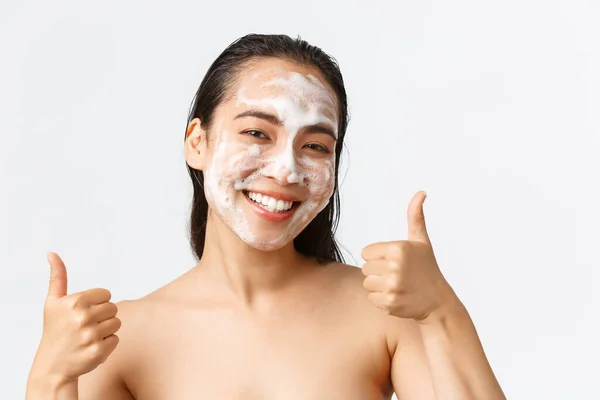 Skincare, kecantikan wanita, kebersihan dan konsep perawatan pribadi. Close-up of satisful happy, smilasian woman standing naked and showing thumbs-up while using cleansing busa on face — Stok Foto