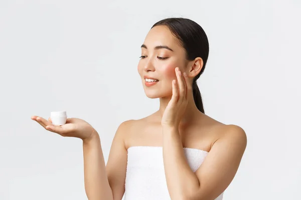 Kecantikan, perawatan pribadi, salon spa dan konsep perawatan kulit. Close-up of beautiful young asian woman standing naked in bath handuk, holding cream on palm and applying it skin, rubbing into face — Stok Foto