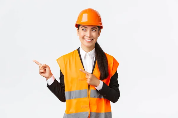 Bouw, bouw en industrieel concept. Glimlachend vertrouwen aziatische vrouwelijke chief engineer in veiligheid helm en reflecterende kleding wijzende vingers linksboven hoek en kijken tevreden — Stockfoto