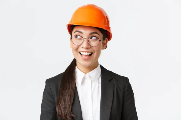 Close-up of creative asian female architect having interesting idea in mind, construction engineer in business suit and safety helmet looking left with pleased smile, standing white background — Stock Photo, Image