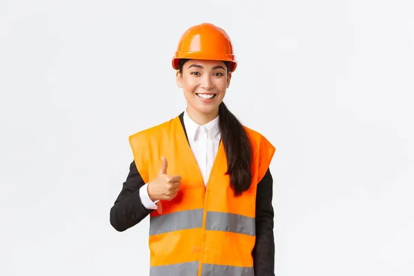Уверенная в себе довольная азиатка-менеджер по строительству в защитном шлеме, светоотражающая куртка, показывающая большой палец вверх в одобрении, поддержке и как работа сотрудников, стоя на белом фоне — стоковое фото