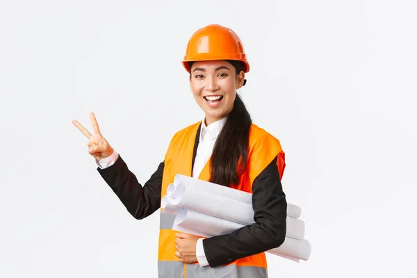 Glimlachend zelfverzekerd aziatische vrouwelijke ingenieur, bouwmanager in veiligheidshelm, dragen blauwdrukken, met twee vingers, zorgen voor het gebouw klaar in de tijd, staande witte achtergrond upbeat — Stockfoto
