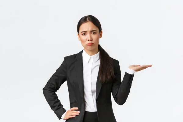 Disappointed and gloomy female sales manager, real estate agent in black suit having bad day, pouting and look upset while showing something, holding hand to the right, white background — Stock Photo, Image