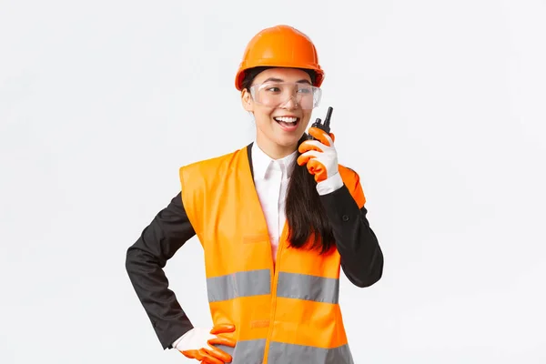 Sebevědomé potěšené asijské ženské inženýrky v bezpečnostní přilbě a uniformě mluví s hlavním architektem pomocí vysílačky. Spokojenost stavebního technika kontaktního týmu pomocí radiotelefonu — Stock fotografie