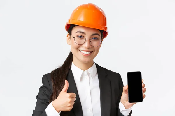 Κοντινό πλάνο της επαγγελματικής χαμογελαστή γυναίκα μηχανικός, διευθυντής κατασκευών στο κοστούμι των επιχειρήσεων και κράνος ασφαλείας, δείχνει οθόνη smartphone και αντίχειρες-up στην έγκριση, λευκό φόντο — Φωτογραφία Αρχείου