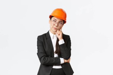 Düşünceli, yaratıcı, Asyalı kadın mimar, inşaat mühendisi, güvenlik kaskı ve takım elbise giyiyor, bina için en iyi seçimi düşünüyor, beyaz arka planda duruyor.
