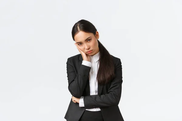 Annoyed and exhausted asian saleswoman looking unamused and bothered from under forehead, standing in black suit, leaning on palm and stare pissed-off, standing white background — Stockfoto