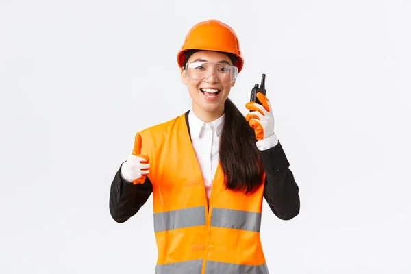 Satisfied happy smiling asian female engineer, industrial technician in safety helmet and uniform showing thumbs-up while praising great work using walkie-talkie, give permission to work — Stock Photo, Image