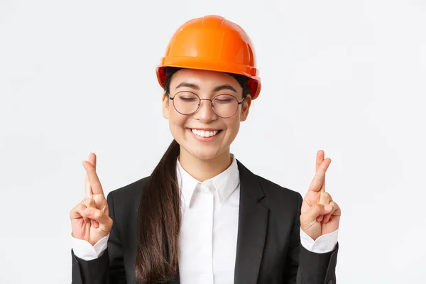 Close-up van hoopvolle optimistische Aziatische vrouwelijke ingenieur, architect die vertrouwen heeft in het project, het dragen van een veiligheidshelm en business suit in afwachting van goede resultaten, kruisvingers veel geluk, witte achtergrond — Stockfoto