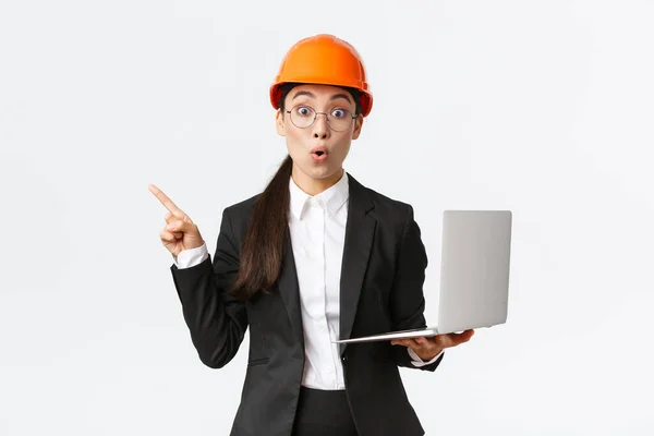 Impresionada mujer empresaria asiática en la fábrica con casco de seguridad y traje de negocios, apuntando con el dedo hacia la izquierda y sosteniendo el ordenador portátil, mostrando el diagrama en la empresa, fondo blanco — Foto de Stock
