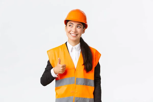 Nöjd glad leende kvinnlig asiatisk arkitekt nöjd med byggnadsarbete, stående i skyddshjälm och reflekterande jacka, tittar övre vänstra hörnet och visar tummen upp i godkännande — Stockfoto