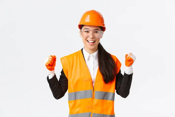 Успешная победительница женщина азиатский инженер, строительный архитектор в защитных очках, перчатки и шлем торжествует, кулак насоса радоваться и сказать да, достичь цели, белый фон — стоковое фото