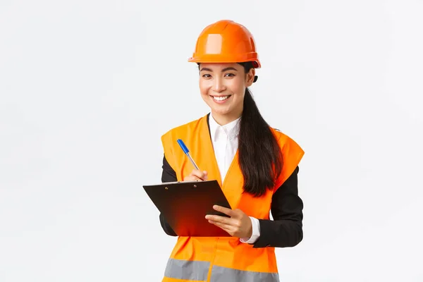 Smiling satisfied asian female construction engineer leading inspection at enterprise, wearing safety helmet and reflective jacket, writing down notes and looking pleased result, white background — Stock Photo, Image