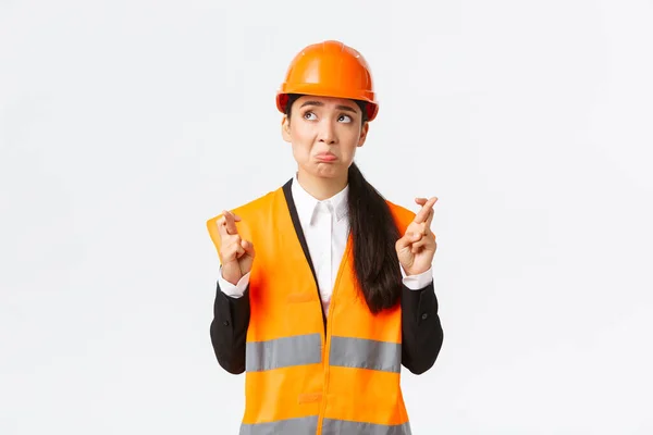 Sad nervous and unconfident asian female construction manager in safety helmet, pouting and look in panic upper left corner, making wish, cross fingers good luck, pleading over white background — Stock Photo, Image