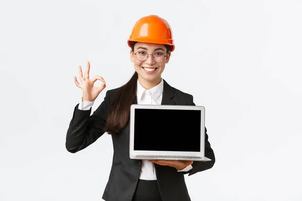 Χαμογελώντας επαγγελματική γυναίκα αρχιτέκτονα, διευθυντής κατασκευών στο εργοστάσιο δείχνει γράφημα, θετικό διάγραμμα, κάνει εντάξει χειρονομία στην έγκριση και κρατώντας το laptop που αντιμετωπίζει οθόνη, φορούν κράνος ασφαλείας — Φωτογραφία Αρχείου