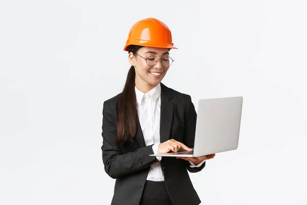 Lächelnde erfolgreiche asiatische Wirtschaftsingenieurin, Fabrikmanagerin in Schutzhelm und Businessanzug mit Laptop, Überprüfung mit Projekt- oder Bauplänen auf dem Bildschirm — Stockfoto