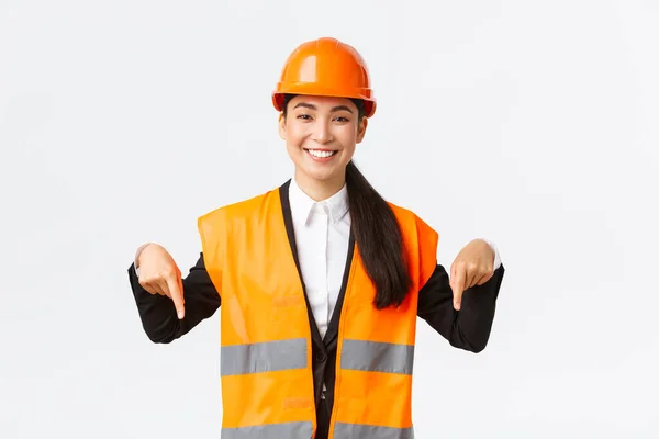 Улыбающийся веселый строительный менеджер, азиатка-инженер в защитном шлеме и отражающая одежда, показывающая объявление. Менеджер проекта женщина указывает вниз, стоя на белом фоне — стоковое фото