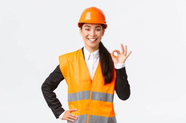 Güvenlik kaskı takmış kendine güvenen profesyonel Asyalı kadın mimar kaliteyi ve işi zamanında temin eder, iyi bir hareket gösterir ve kararlı gülümser, iddialı durur, bir şeyi garantiler ve garantiler.